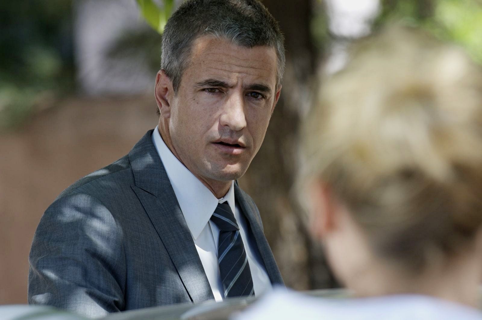 Man in a suit looking intently at someone off-camera. He has graying hair and a concerned expression, outdoors.