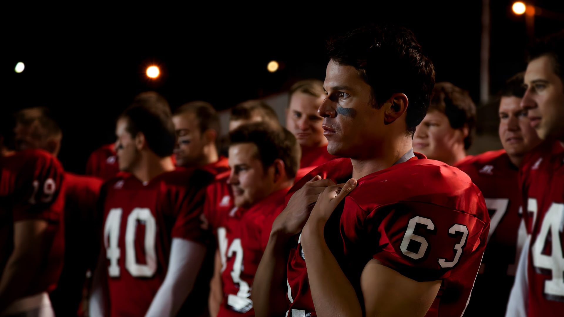 A football team in red uniforms stand on a field at night. A player with the number 63 looks thoughtful.