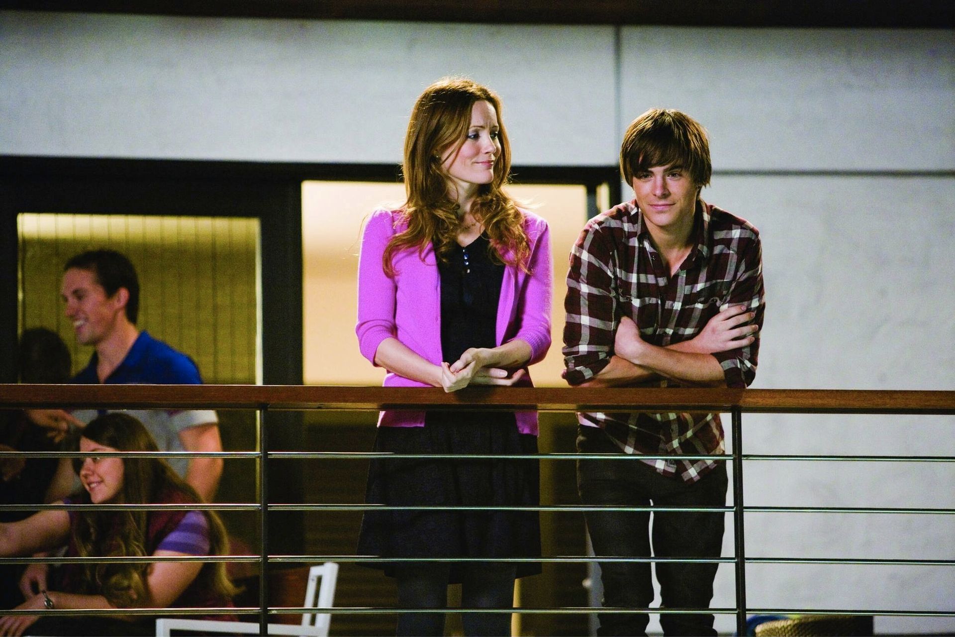 A young woman in a pink blazer and a young man in a plaid shirt smile together on a balcony, observing an event.