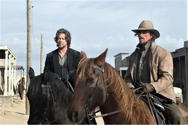 Two men on horseback in a dusty Western town; one wears a brown coat and hat, the other a dark coat.