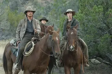 Three people on horseback in a rugged, outdoor setting. A man in a hat and jacket leads, followed by a child and another man.
