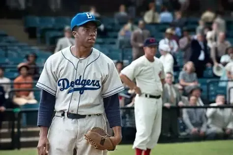 Chadwick Boseman as Jackie Robinson in a Dodgers uniform, looking focused on a baseball field. An opposing player stands nearby.