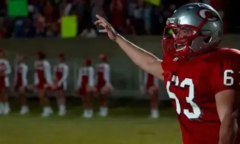 Football player in a red uniform and silver helmet with the number 53 on the field, celebrating with his arm raised. Cheerleaders in the background.