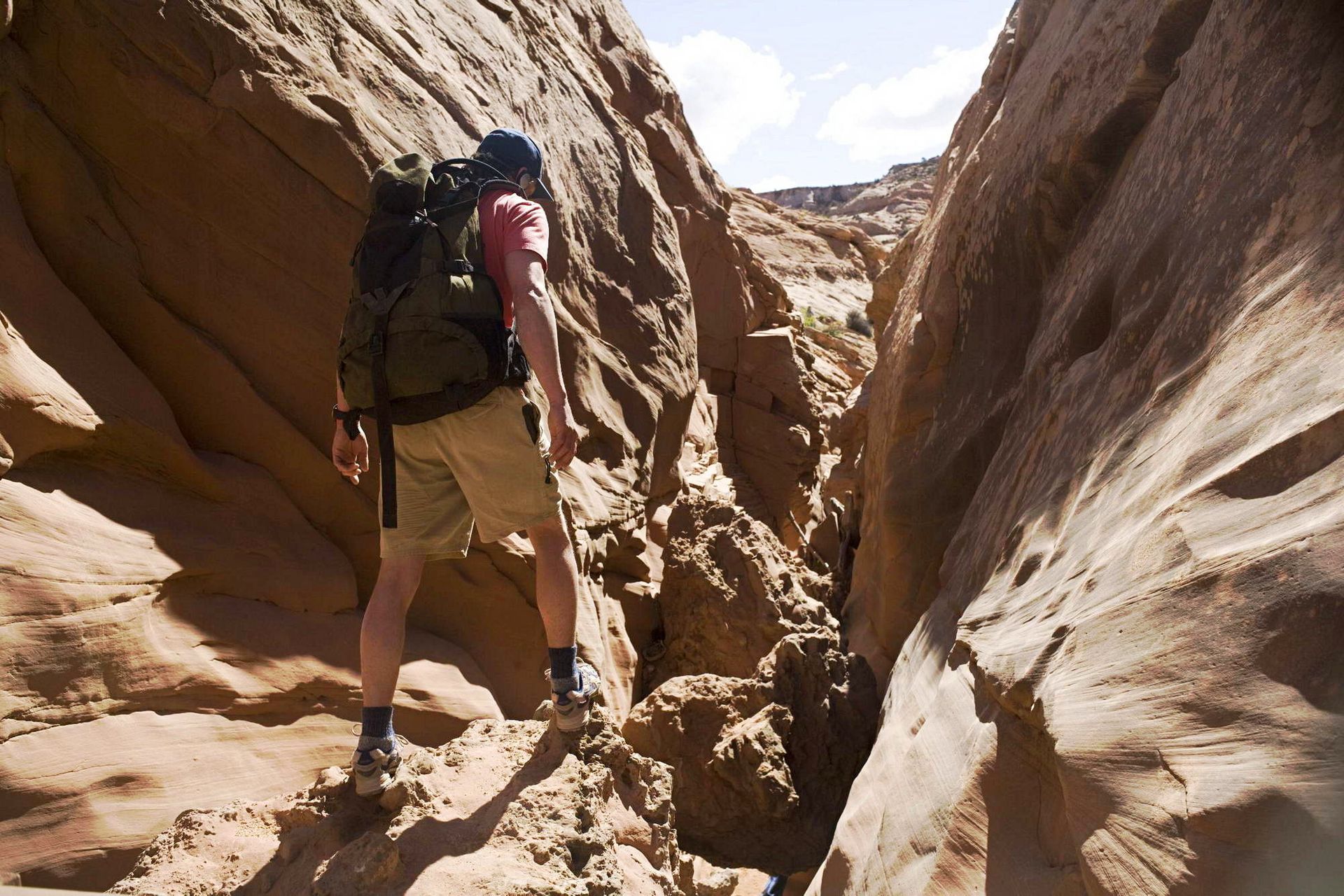 A hiker in shorts and backpack traverses a narrow canyon. The red rock walls create a sunlit path.