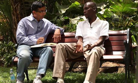 Two men sit on a park bench, one interviewing the other. The interviewer wears a blue shirt and glasses, while the interviewee wears a white shirt and khaki pants.
