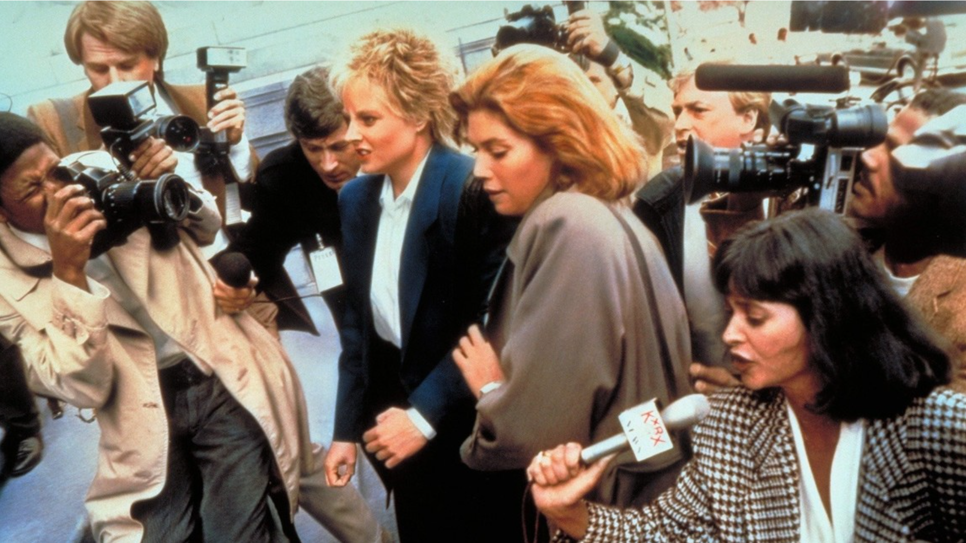 Two women surrounded by paparazzi, walking outside. One woman in a blue suit, the other in a brown coat.