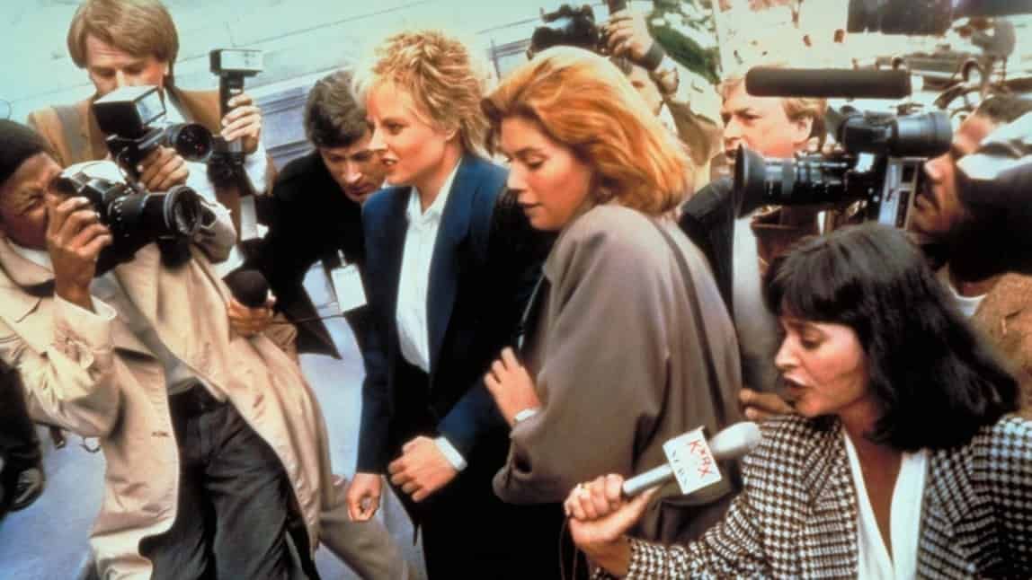 Women surrounded by paparazzi, one holding a microphone, all appearing at an event.