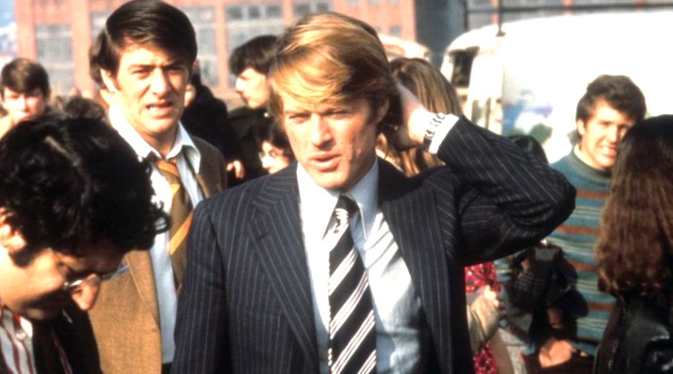 Michael Douglas in a convertible car, wearing sunglasses and a suit, looking to the side.