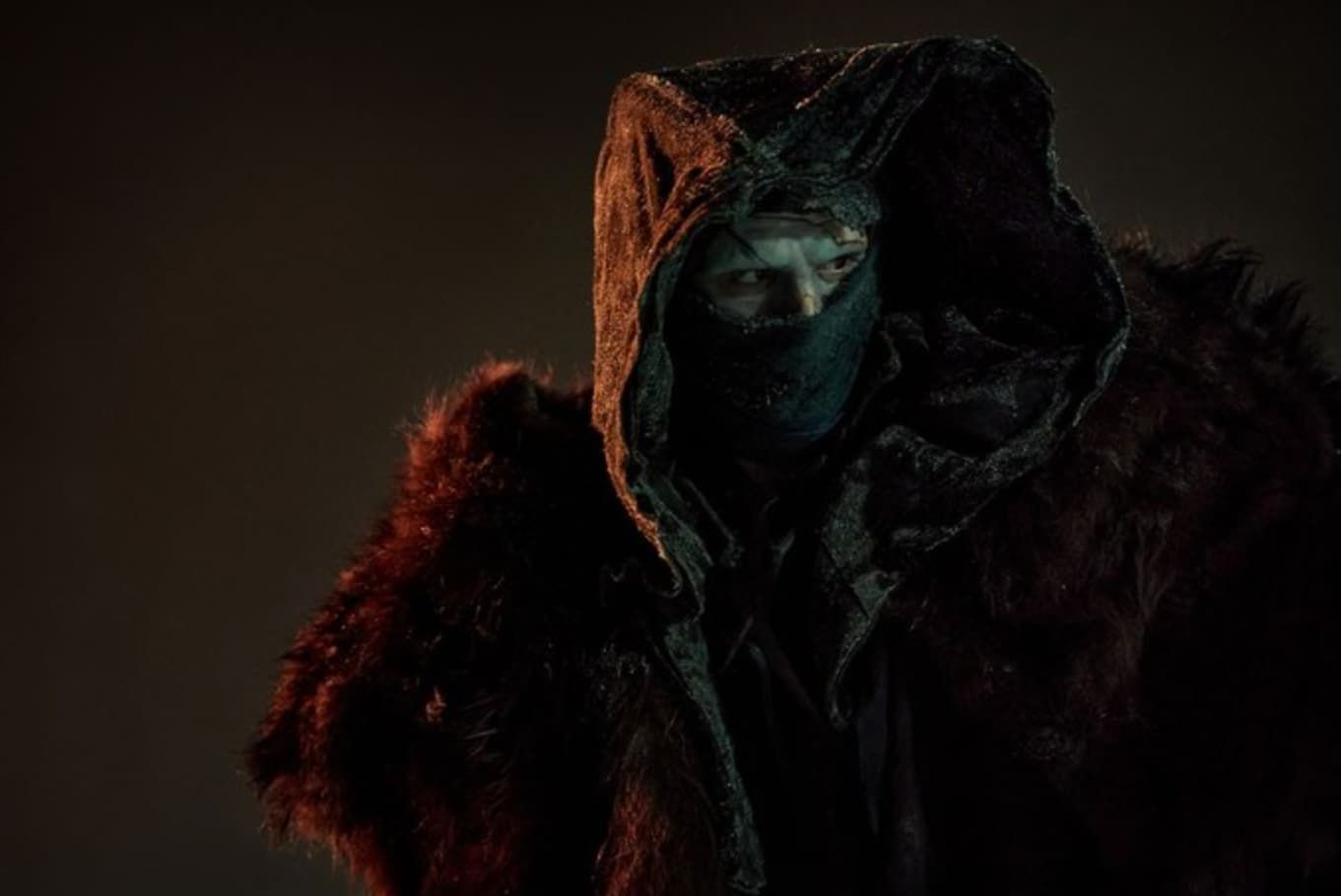 Frankenstein in dark hooded coat, face mostly covered by a scarf. Fur trim, dim setting.