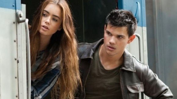 Lily Collins and Taylor Lautner on a train, looking concerned. Collins has long brown hair and is leaning out the door. Lautner wears a leather jacket.