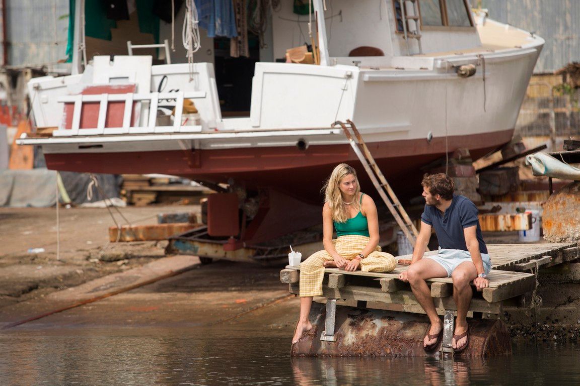 A blonde woman and a man sit on a dock by a boat. She wears a green top and pants. He wears a blue shirt and shorts.