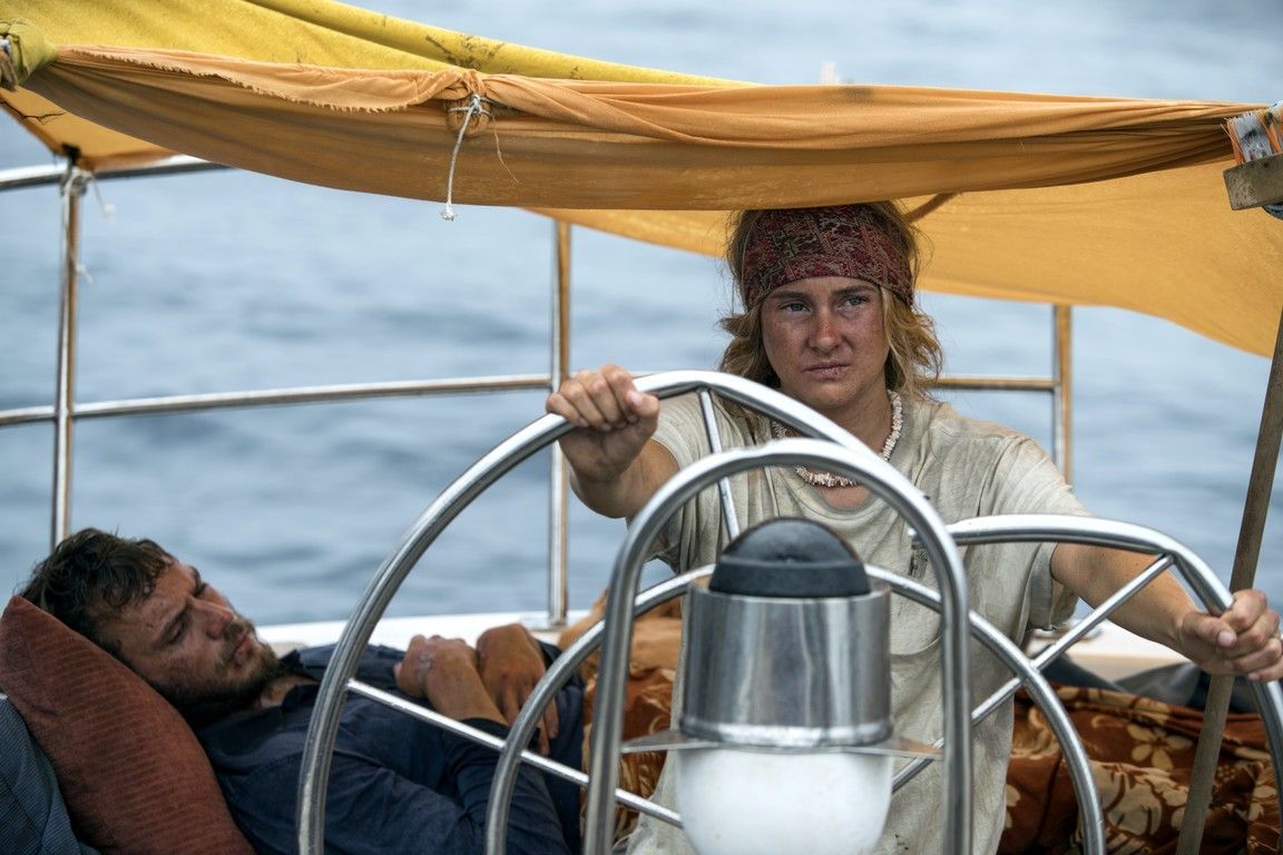 A woman wearing a headscarf steers a sailboat, while a man sleeps nearby. The boat is on the water under a yellow canopy.