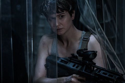 Woman with short hair, holding a rifle, looks at the camera through a reflective glass, dimly lit setting.