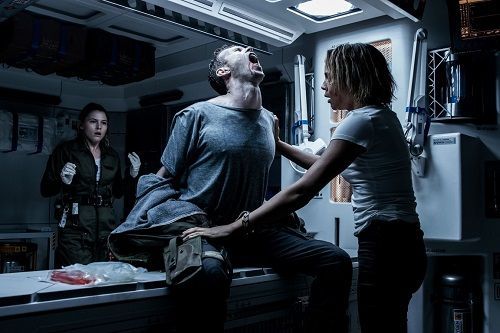 Man clutches his neck, writhing in pain, as a woman tries to help. Another woman watches in horror in a dimly lit, sterile environment.