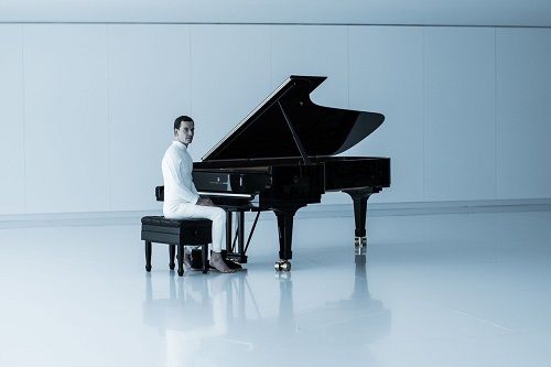 A man in white attire sits at a black grand piano in a bright, minimalist room. He is positioned as if ready to play.