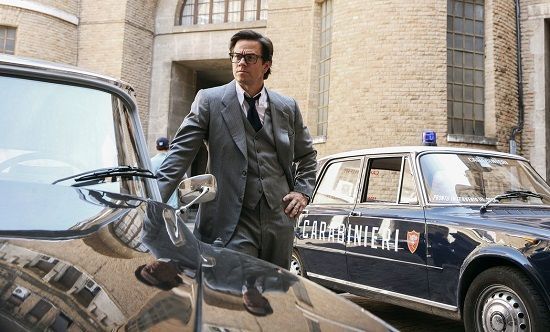 Man in gray suit stands near vintage police car in front of a building.