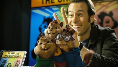 A man smiles, holding chipmunk puppets. The chipmunks are dressed in colorful tops, and one wears glasses. Album art is in the background.