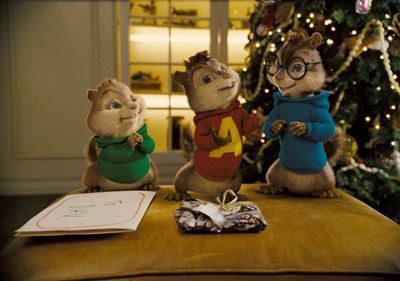 The Chipmunks stand around a gift on a table in front of a Christmas tree. Alvin in red looks excited, while Simon in blue smiles. Theodore in green smiles shyly.