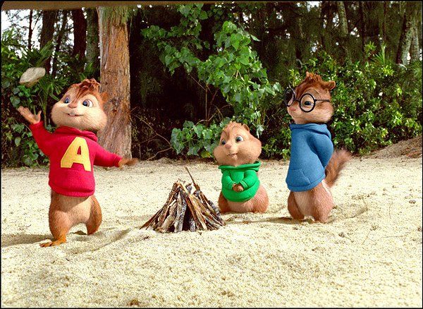 Alvin, Simon, and Theodore chipmunks gathered around a small campfire on a sandy beach. Alvin gestures with raised arms.