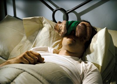 Man asleep in bed with a sock over his eyes. The sock is green and tan, the bedding is white.