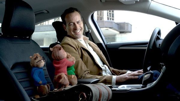 A man smiles while driving, with Alvin and Simon chipmunk dolls on the front seat. The scene is in a car with an urban background.