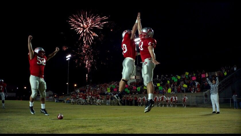 Football players in red uniforms celebrate a win on the field under fireworks. Two jump in the air for a high five, as others cheer.