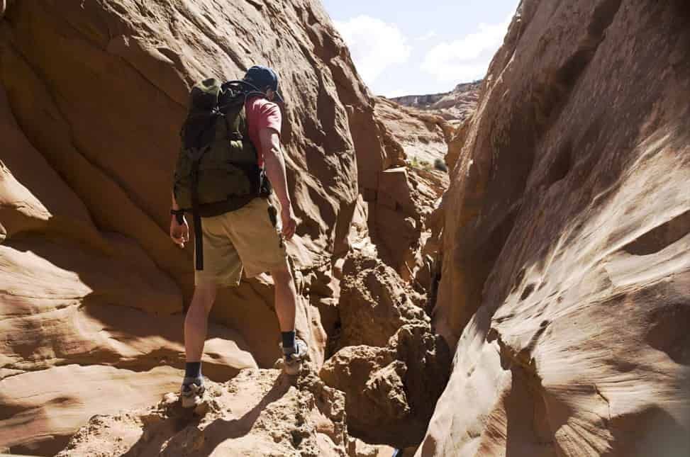 Hiker with backpack traversing a narrow canyon carved in reddish rock under a bright sky.