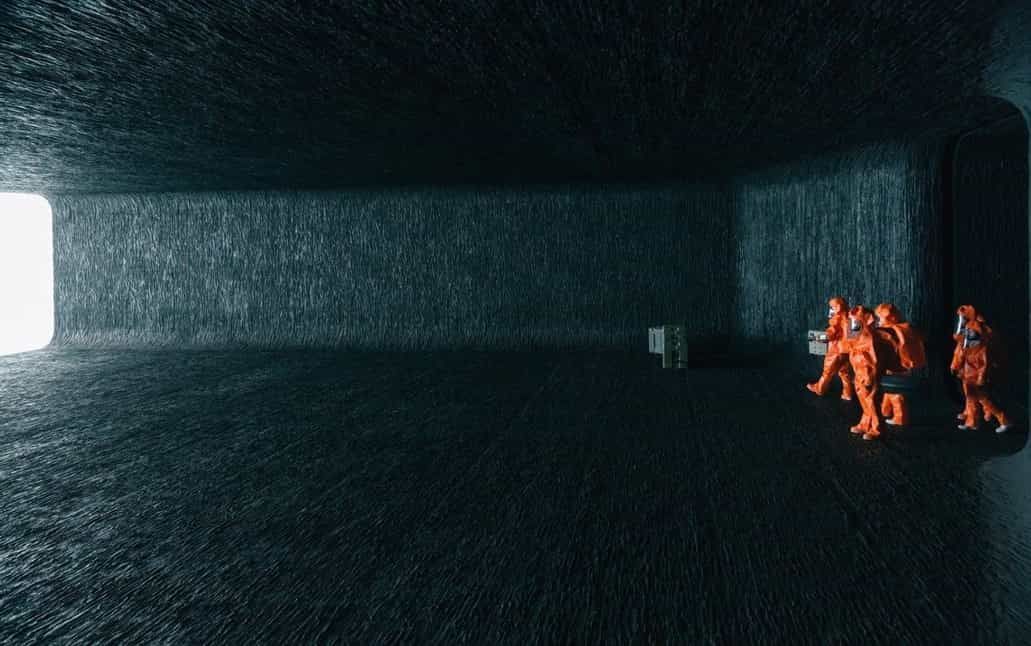 Orange-suited figures stand in a dark, textured room near a light-filled opening.