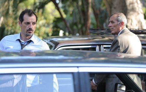Two men near cars; one in a suit, the other in a light blue shirt with a loosened tie.