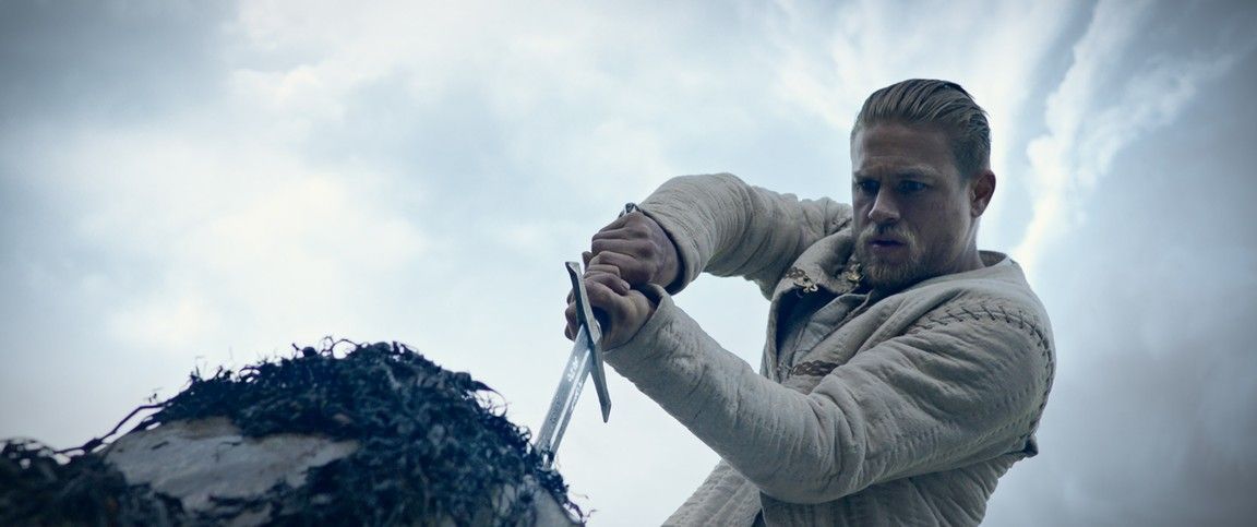 Man in a white coat, focused, plunging a sword into a rock in a cloudy environment.