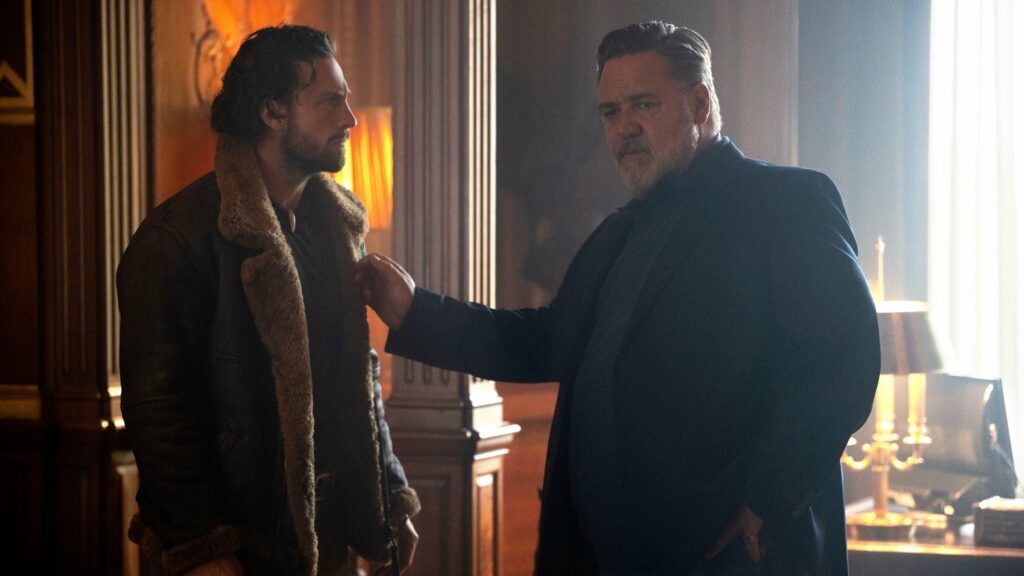 Man in brown jacket facing older man in black coat, interior setting. Older man touches the younger's jacket.