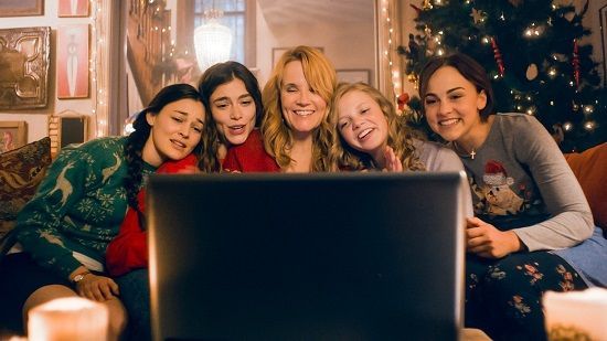 Five people smiling at a laptop screen in a cozy room decorated for Christmas.