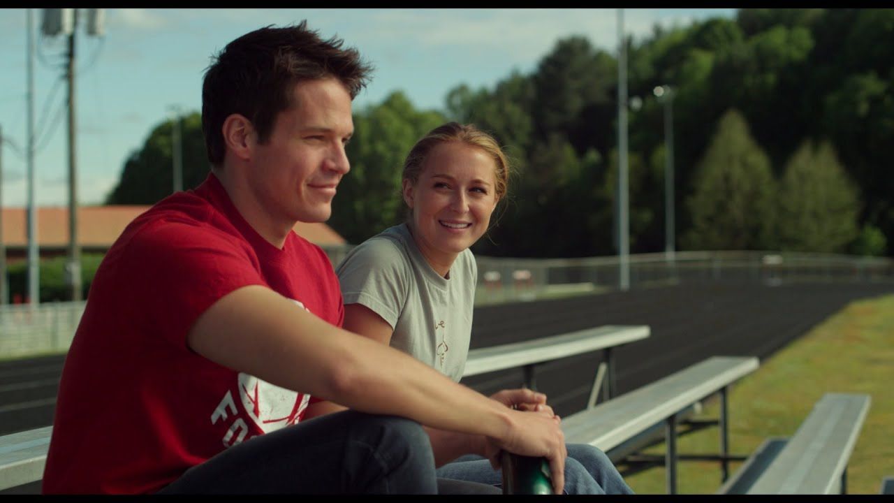 Two people smiling, seated on bleachers at a track. Man in red shirt, woman in gray shirt. Green trees in the background.