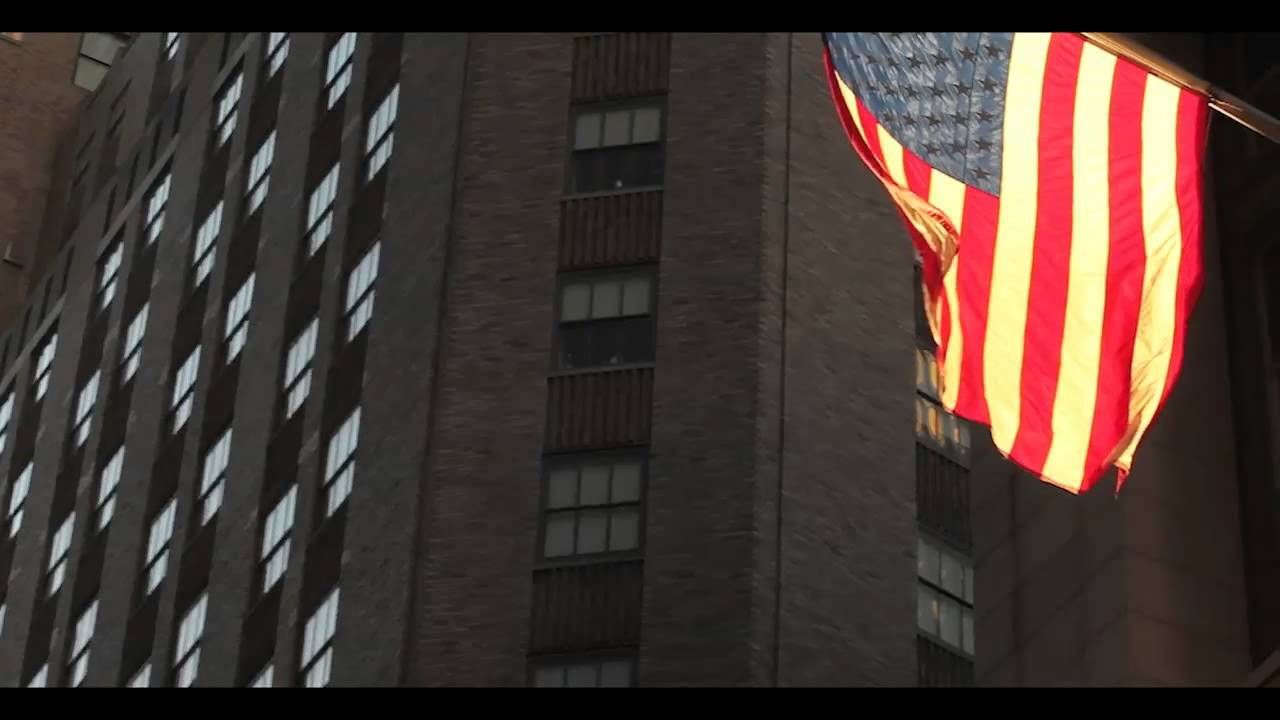 American flag waving in front of a tall brick building with many windows. Sunlight is reflecting off the building's windows.