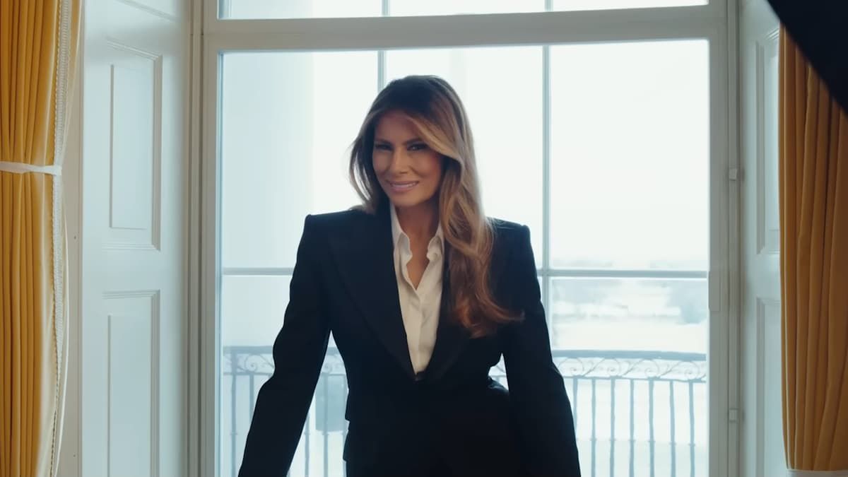 Melania Trump in a black suit smiles, standing near a window with the White House in the background.