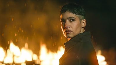 Woman with short, dark hair looks toward the camera. Fire burns in the background, casting an orange glow.