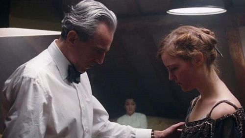 Older man adjusts the dress of a young woman indoors; a woman watches from the background.
