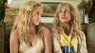 Two blonde women in a truck bed, looking at each other. One wears a tank top, the other a t-shirt and cardigan.
