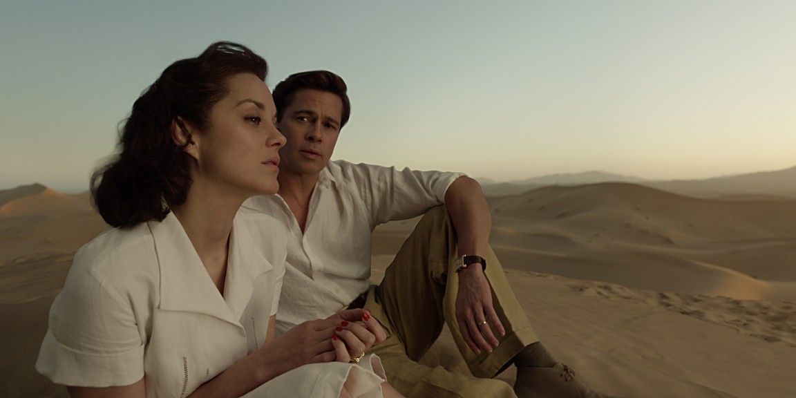 A woman and a man sit on a sand dune, looking out at the desert. The woman wears a white dress, the man a cream shirt and khaki pants.