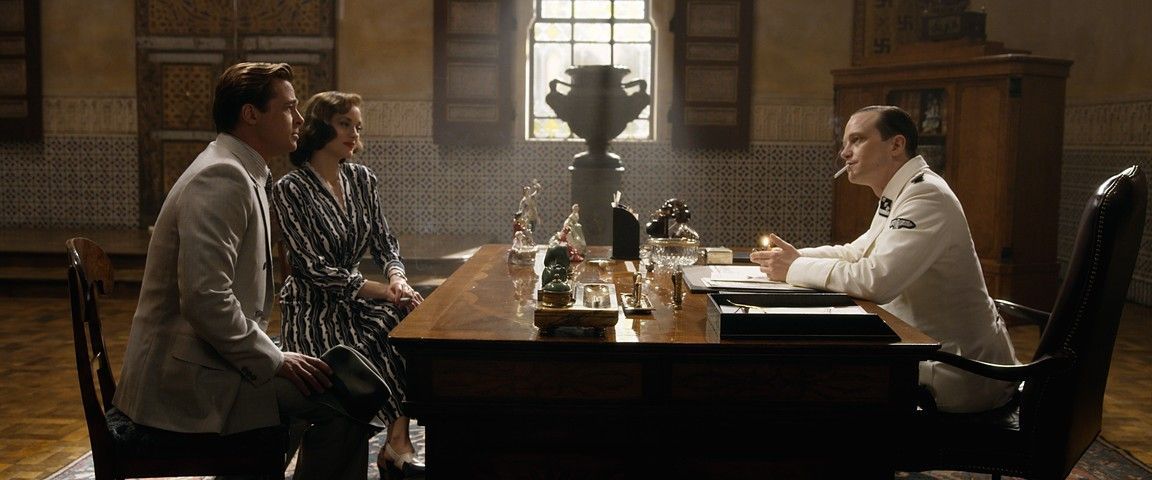 A man and woman sit across from a doctor in a well-lit room. The doctor, in a white coat, gestures while smoking a cigarette.