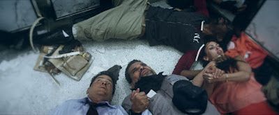 People lying on a surface; some appear injured, one person holds a gun; setting is indoors.