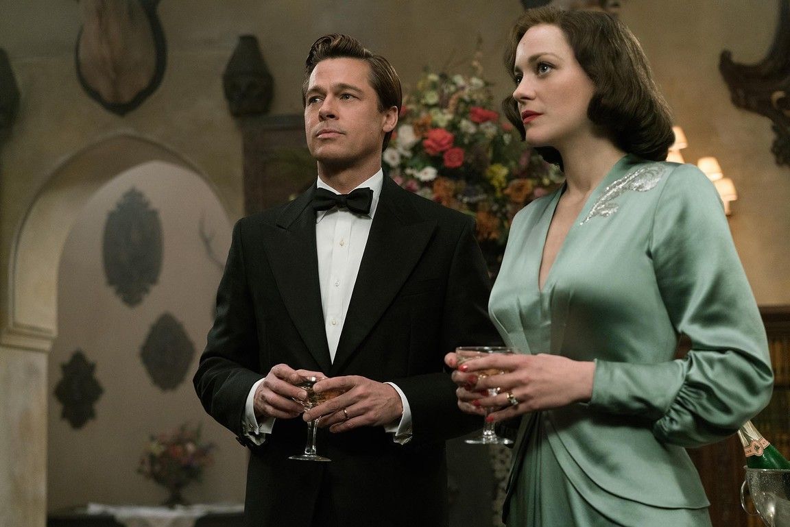 Brad Pitt in a tuxedo and Marion Cotillard in a teal dress stand holding drinks in a room with ornate decor.