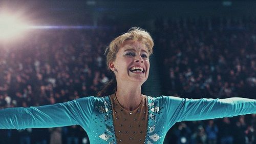 Figure skater, Margot Robbie, smiles brightly with arms outstretched on the ice, crowd in the background.