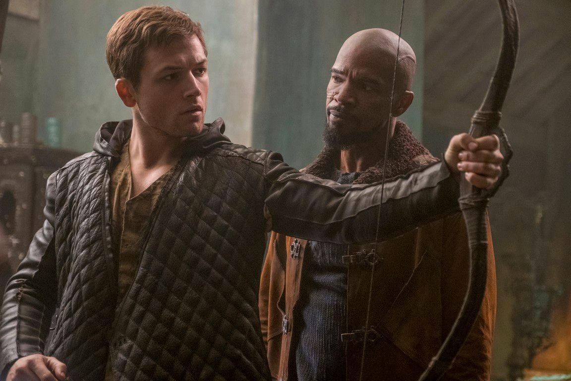 Robin Hood and Little John looking at a bow; indoor setting.