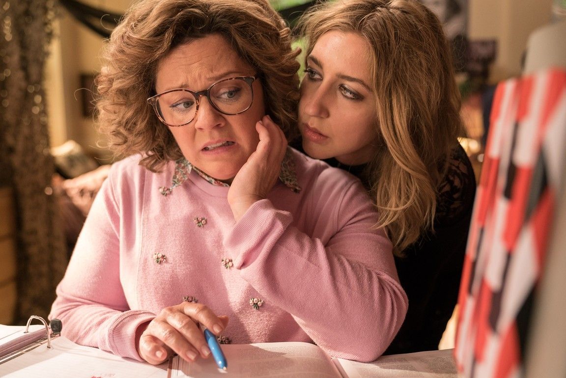 Melissa McCarthy and Jillian Bell in a pink sweater and black shirt, respectively, appearing worried at a table.