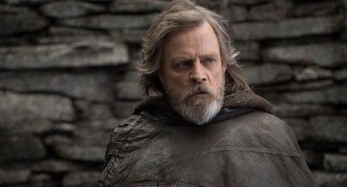 Old Luke Skywalker with a gray beard and hooded cloak, standing in front of a stone wall.