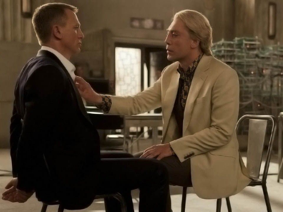 Daniel Craig as James Bond is held captive and facing Javier Bardem as Silva in a dimly lit room. Silva wears a light blazer and touches Bond’s chest.
