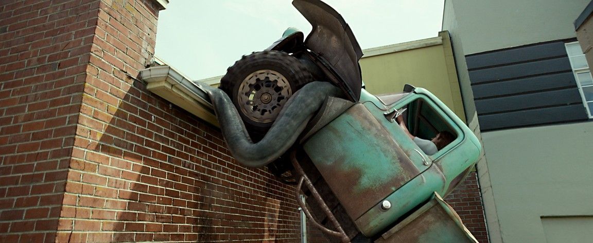 A large snake wrapped around the front of a green pickup truck that is crashed through a brick wall.