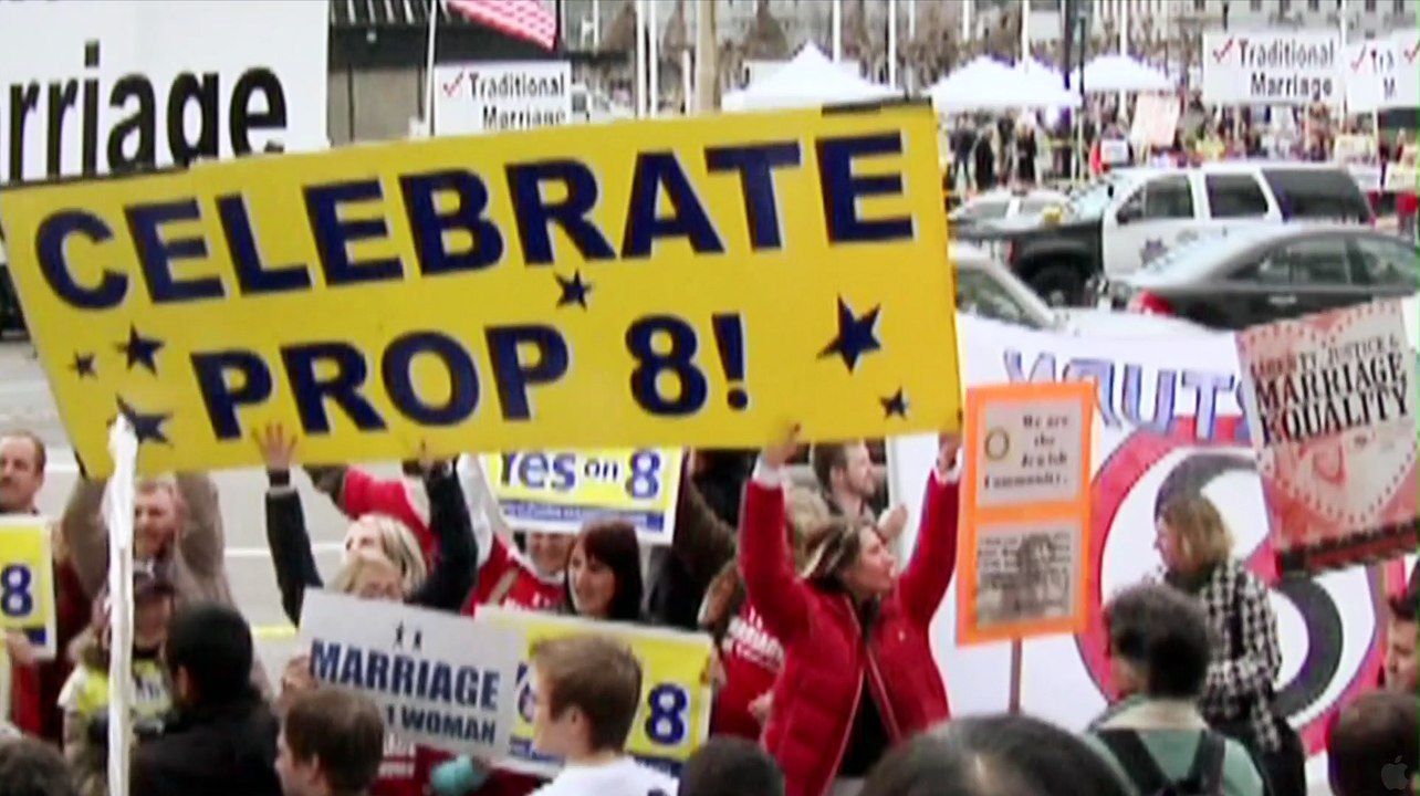 People holding signs celebrating Proposition 8 at a rally. The signs are yellow, blue, and white, and the crowd appears joyful.