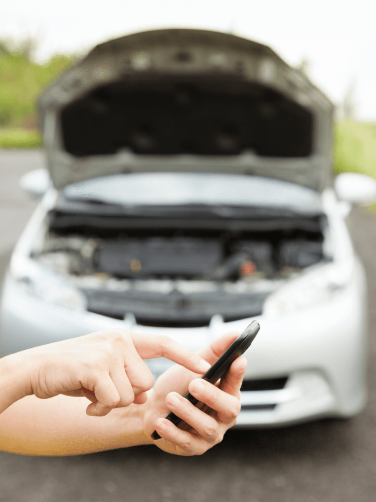 Person using phone, silver car with open hood in the background.